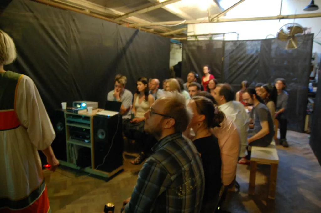 An audience seated in an industrial studio with black fabric walls, watching a presentation beside a projector and large speakers.
