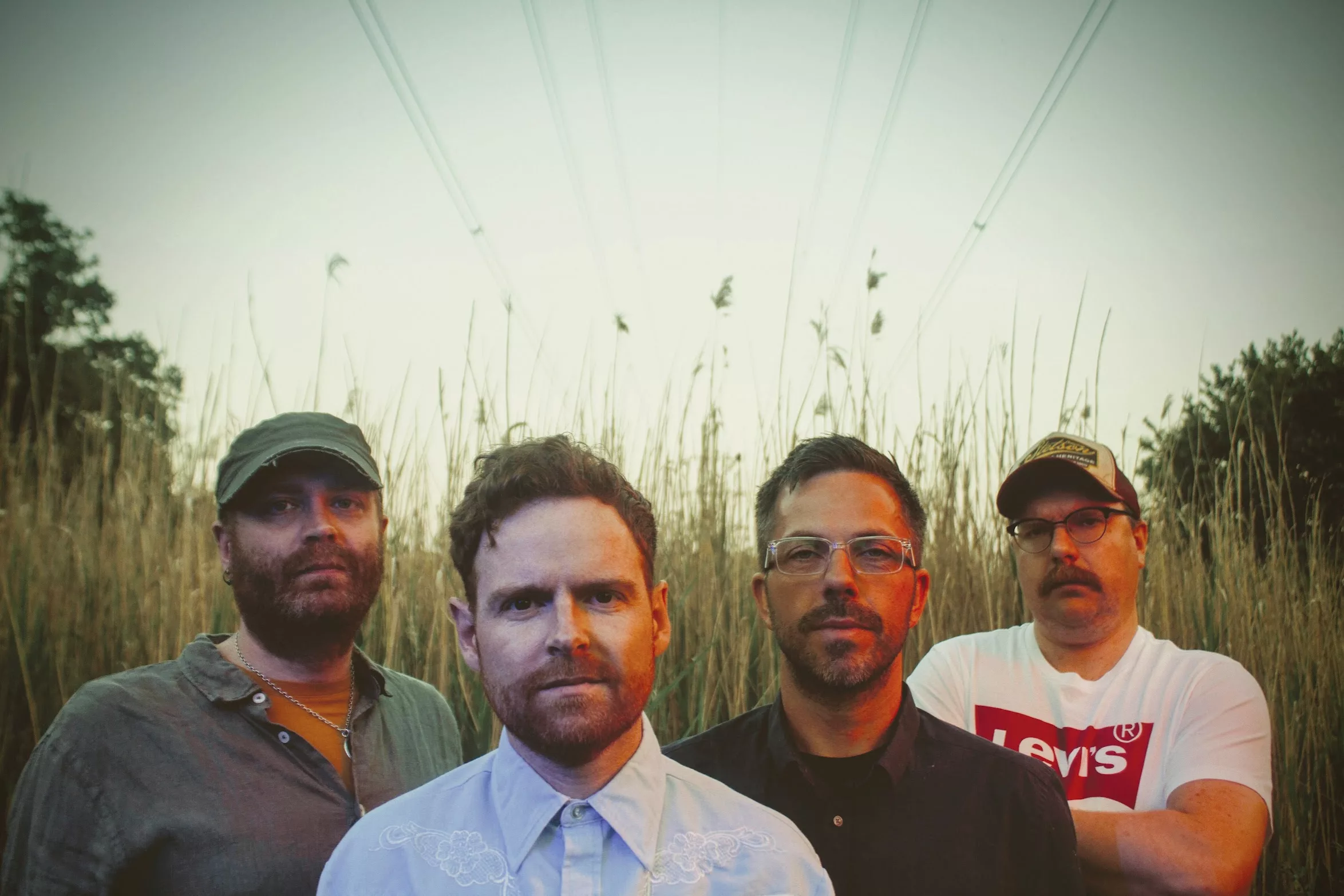 Four men pose in a field of tall dry grass beneath overhead power lines. One wears a light blue western shirt, another a white Leviās t-shirt.