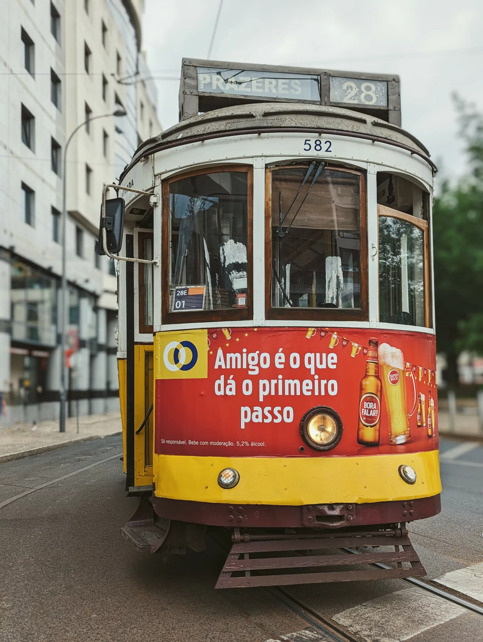 Yellow and white vintage number 28 tram in Lisbon with a large red beer advertisement on its front, stopped on tracks beside city buildings.