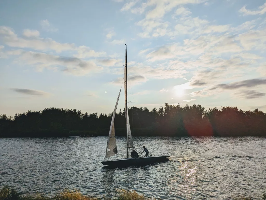 A small sailboat on a shimmering lake at sunset, with two figures on board silhouetted against a wooded shoreline and a cloudy sky.