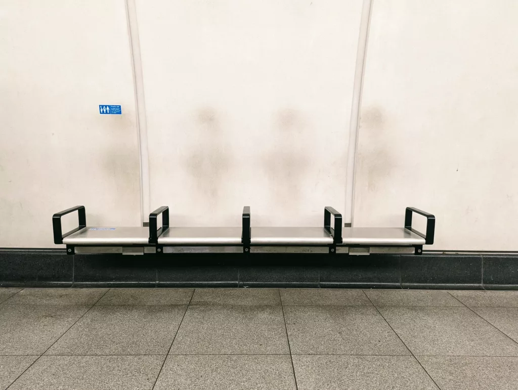 Four metal station seats against a white wall with dark, ghostly smudges where passengers' heads have rested.