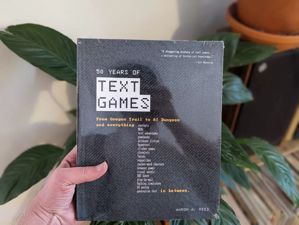 A hand holding the book 50 Years of Text Games by Aaron A. Reed, shrink-wrapped, against a background of large green leaves.