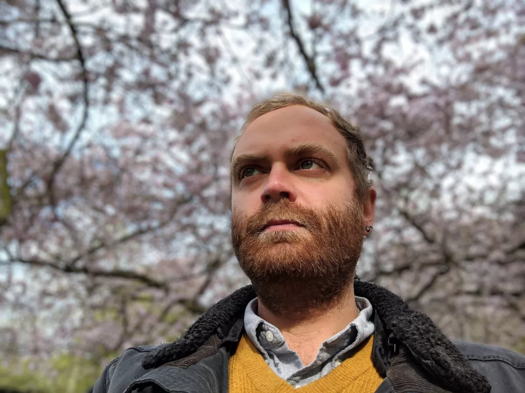 The image shows Jay Springett, also known as @thejaymo, standing outdoors with a thoughtful expression. He is wearing a yellow sweater, a collared shirt, and a jacket with a shearling collar. Behind him, there are trees with branches full of delicate blossoms, suggesting a springtime setting. The soft, natural light highlights his face, while the blurred background of blooming trees adds a serene, contemplative atmosphere.