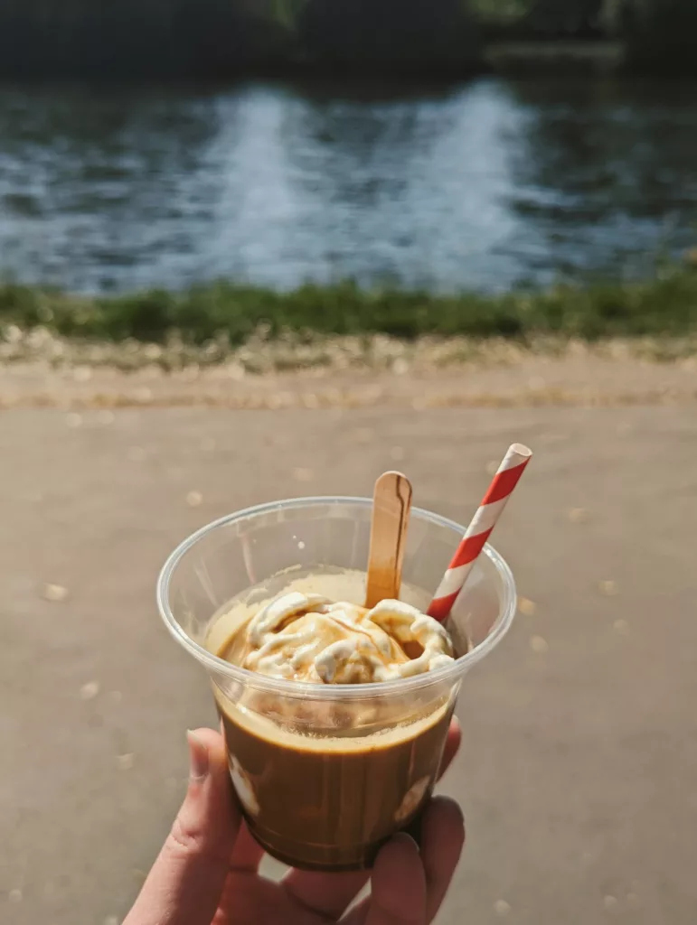 An affogato with whipped cream, a wooden spoon, and a red striped straw in a clear cup, held against a blurred riverside background.