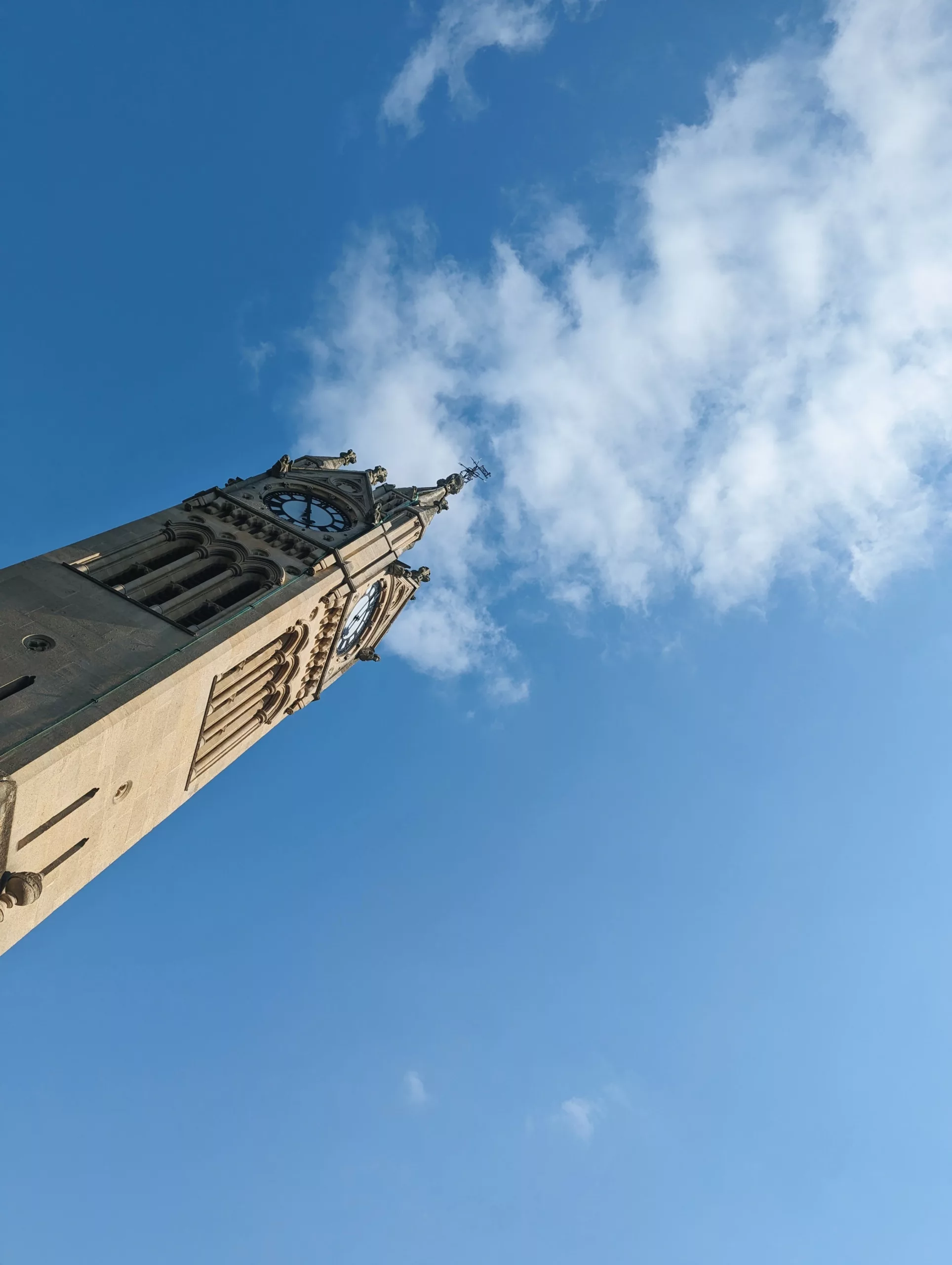 A low-angle view of a stone Gothic clock tower reaching into a bright blue sky with wispy white clouds.