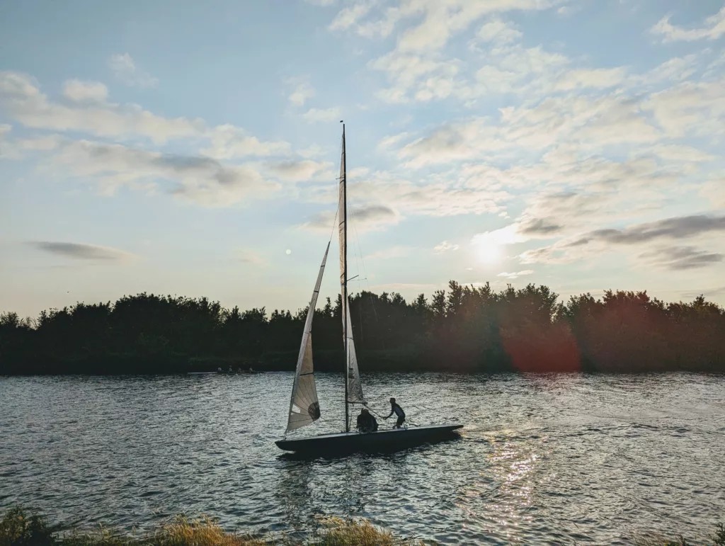 Two people on a sailboat on a river at sunset, silhouetted against a treeline under a vast sky with scattered clouds.