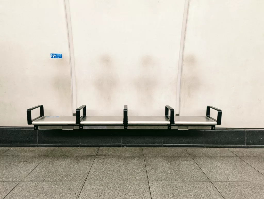 A row of four silver metal seats with black armrests against a white wall marked with faint, vertical grey smudges.