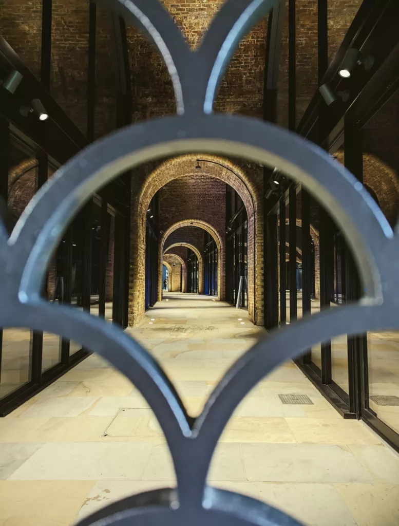 A series of receding brick arches along a stone-paved corridor, framed by the blurred silhouette of a decorative metal gate.