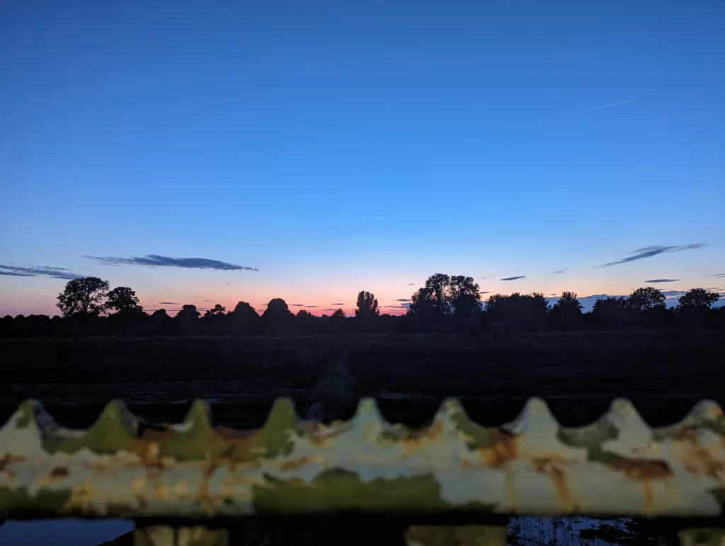 Twilight sky with a pink glow on the horizon over dark trees, viewed over a rusted, jagged metal fence in the foreground.