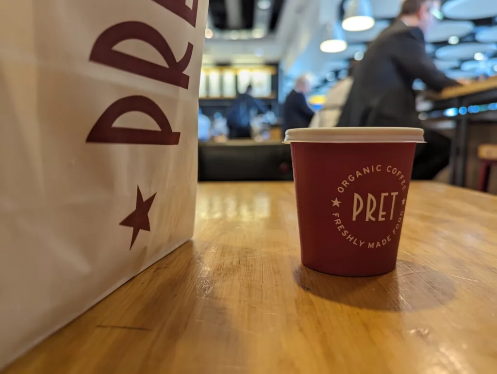 A Pret A Manger paper bag and small coffee cup on a wooden table, with blurred people and lights in a cafe background.