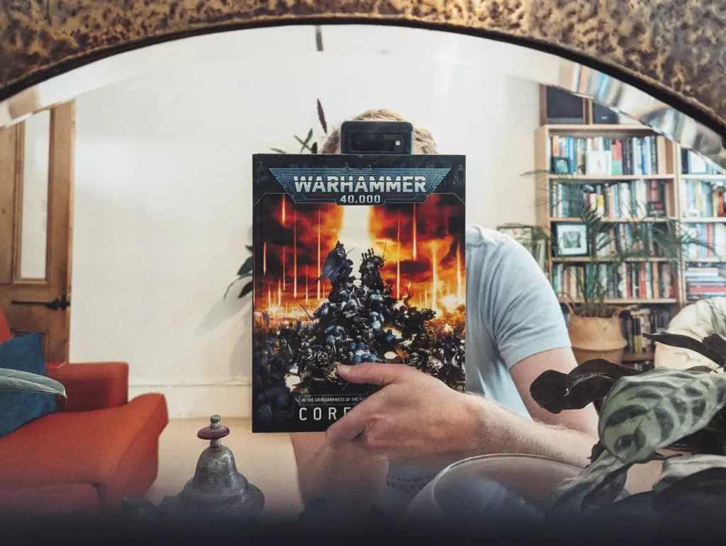 A mirror reflection of a person holding a Warhammer 40,000 Core Book in front of their face, set against a living room with a full bookshelf.