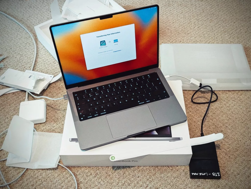 A MacBook Pro sits on its box showing a data transfer screen, surrounded by packaging and an external hard drive on a beige carpet.