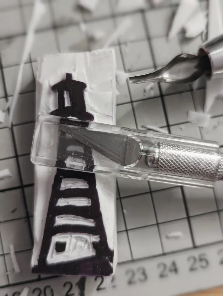 A lighthouse design carved into a white rubber block using a craft knife and gouge on a gridded cutting mat covered in shavings.