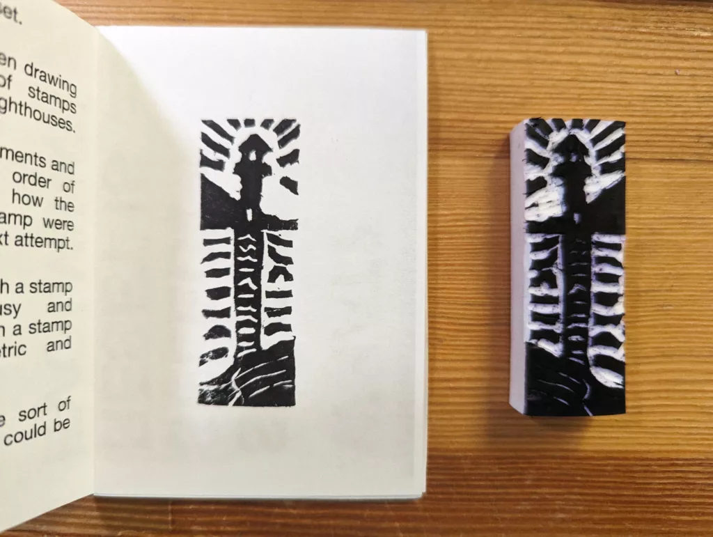 A black ink linocut print of a lighthouse in a small white zine next to the tall, hand-carved stamp on a wooden surface.
