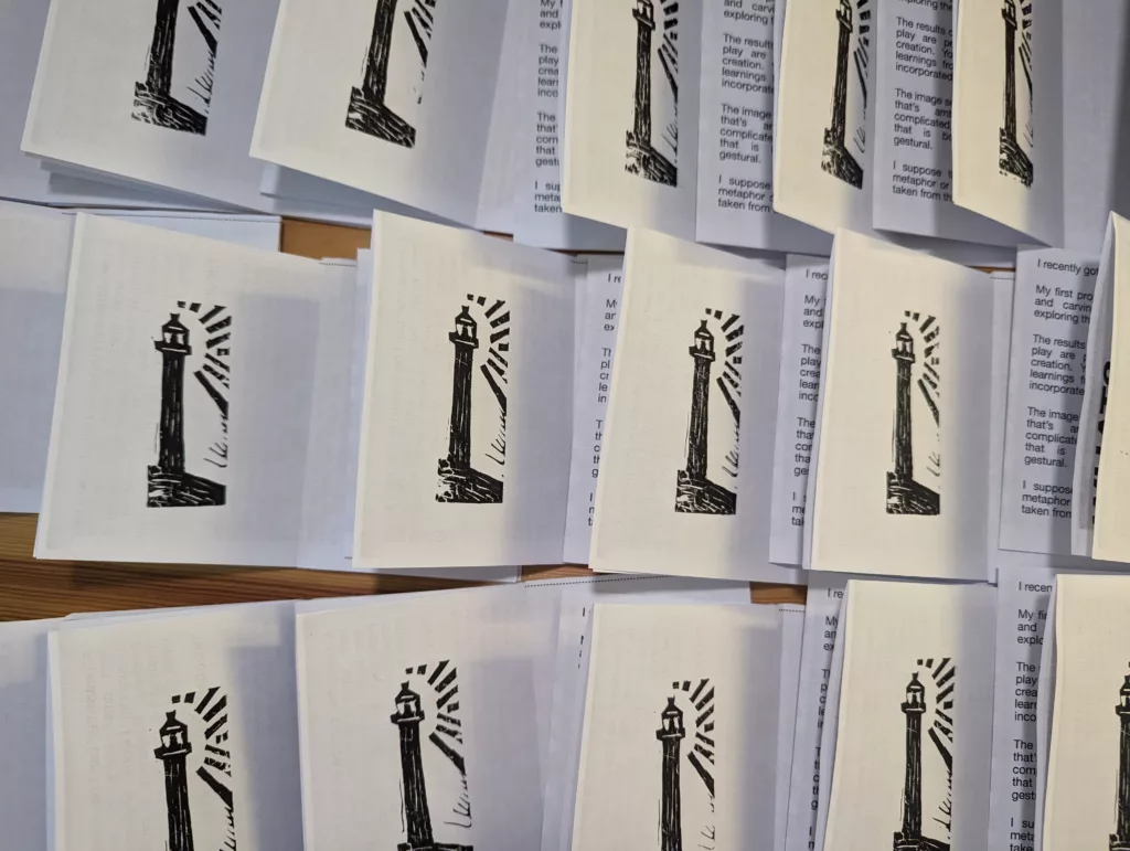 Rows of small white pamphlets featuring a black linocut lighthouse illustration, spread out across a wooden surface.