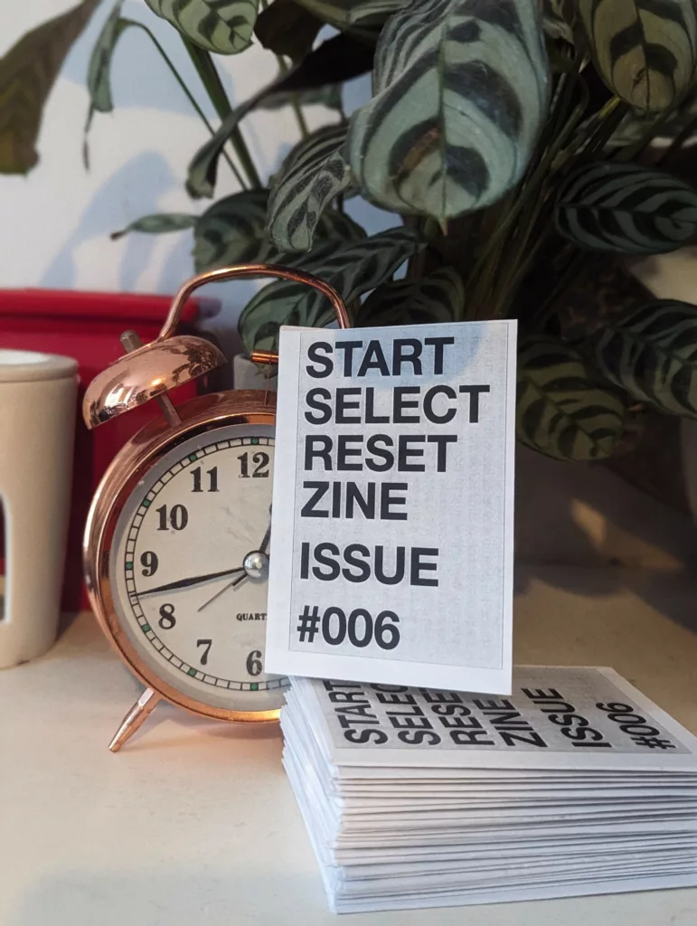 Small white zines titled START SELECT RESET ISSUE #006 leaning against a copper alarm clock beside a leafy green houseplant.