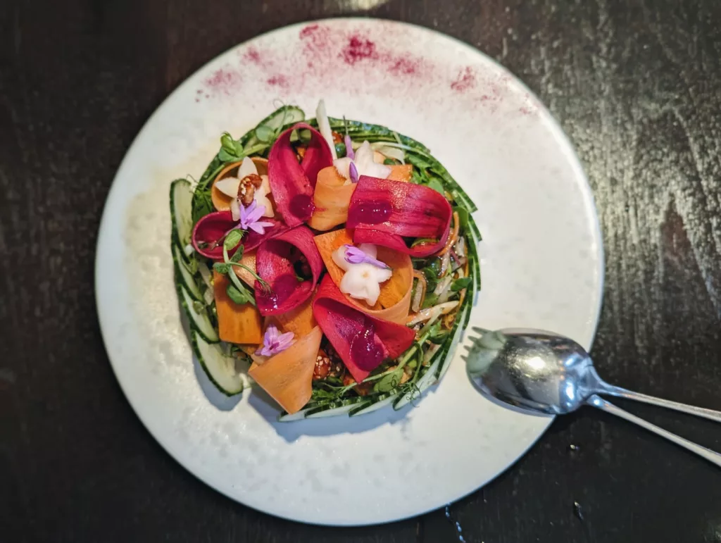A circular salad with a cucumber base, carrot and magenta ribbons, and tiny purple flowers on a white plate dusted with pink powder.