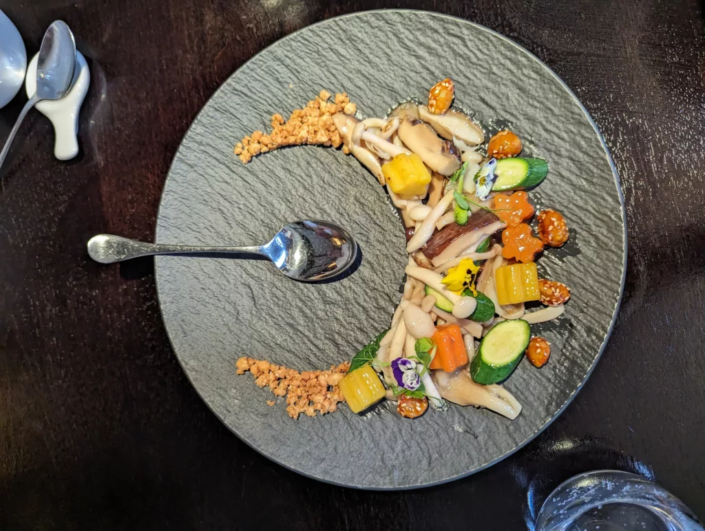 Stir-fried mushrooms, vegetables, and edible flowers arranged in a crescent on a grey textured plate with a silver spoon on a dark wooden table.