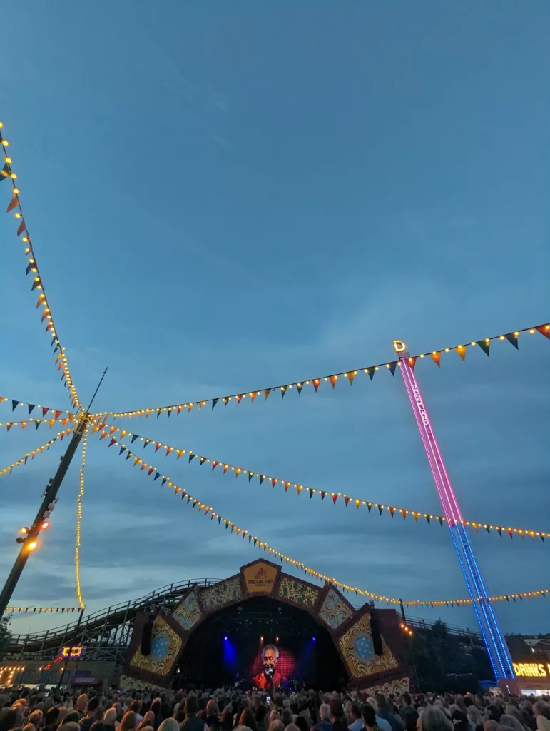 A crowd at a Dreamland Margate concert under festive bunting and a tall neon-lit ride against a deep blue twilight sky.