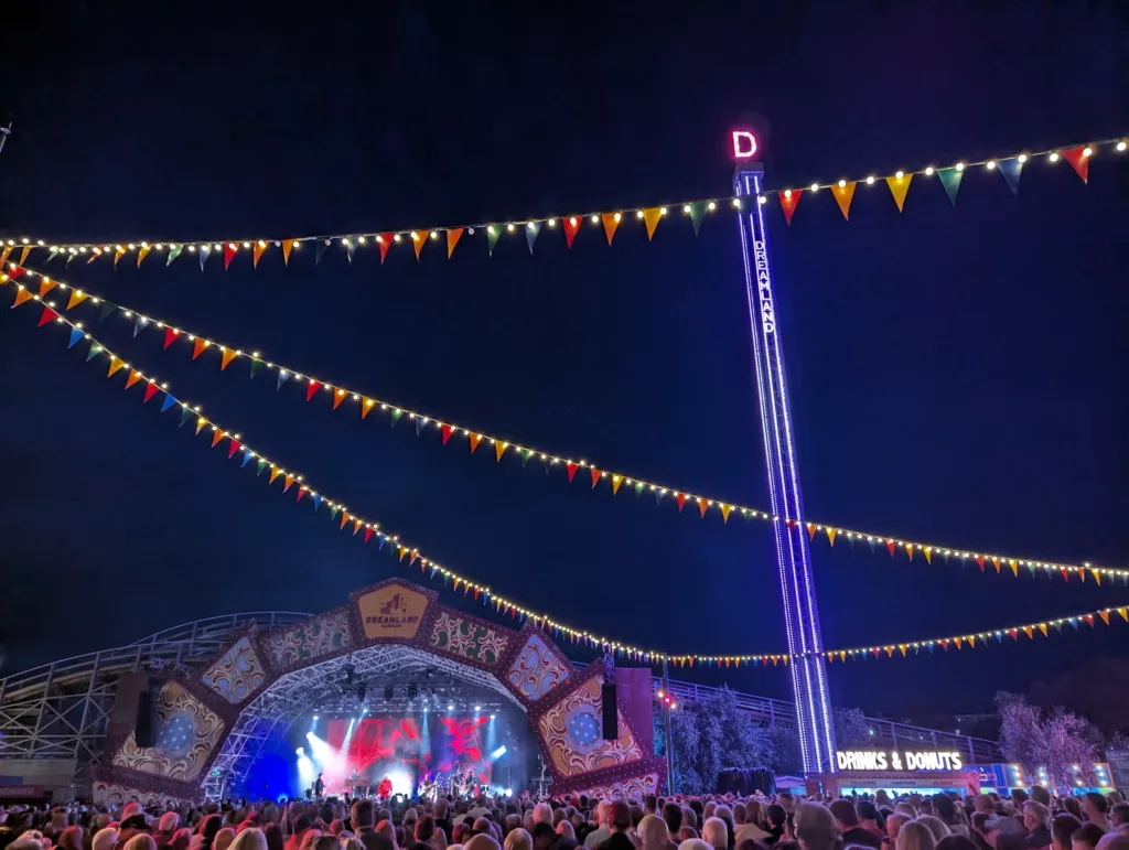 A night concert at Dreamland under strings of bunting lights, featuring an ornate stage, a large crowd, and an illuminated drop tower ride.