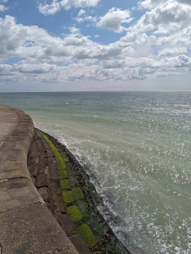Curved stone sea wall with bright green moss on lower steps meeting the pale green ocean under a bright, cloud-filled sky.