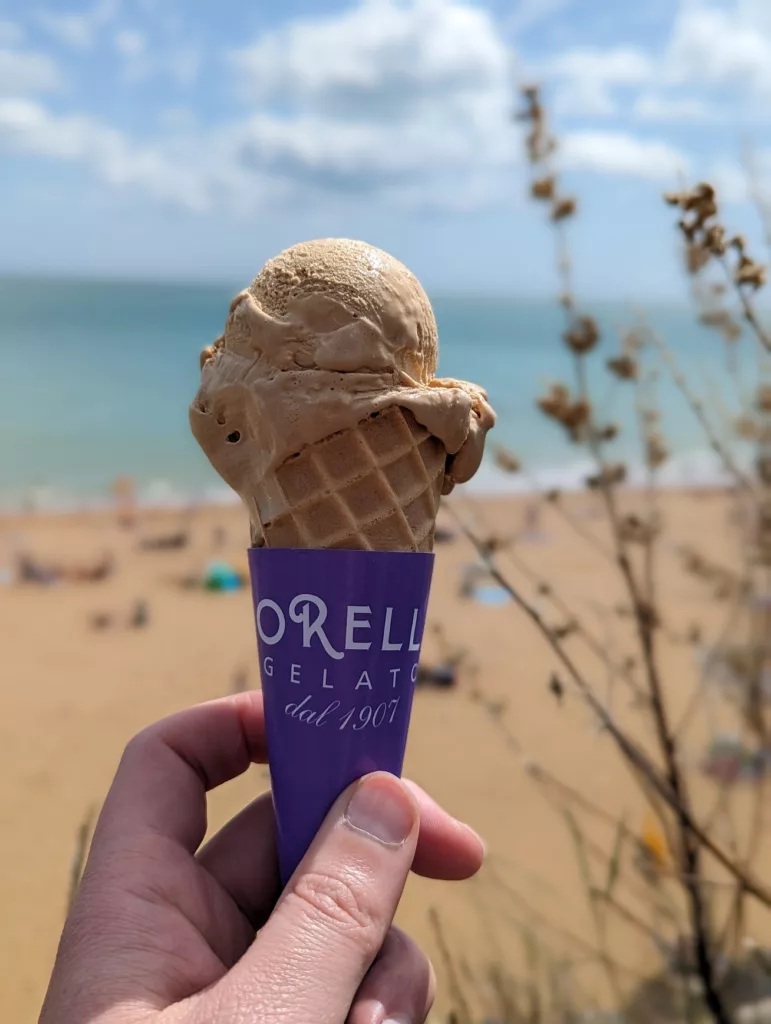 Hand holding a Morelli's gelato cone with a purple wrapper, set against a blurred sunny beach and turquoise sea.