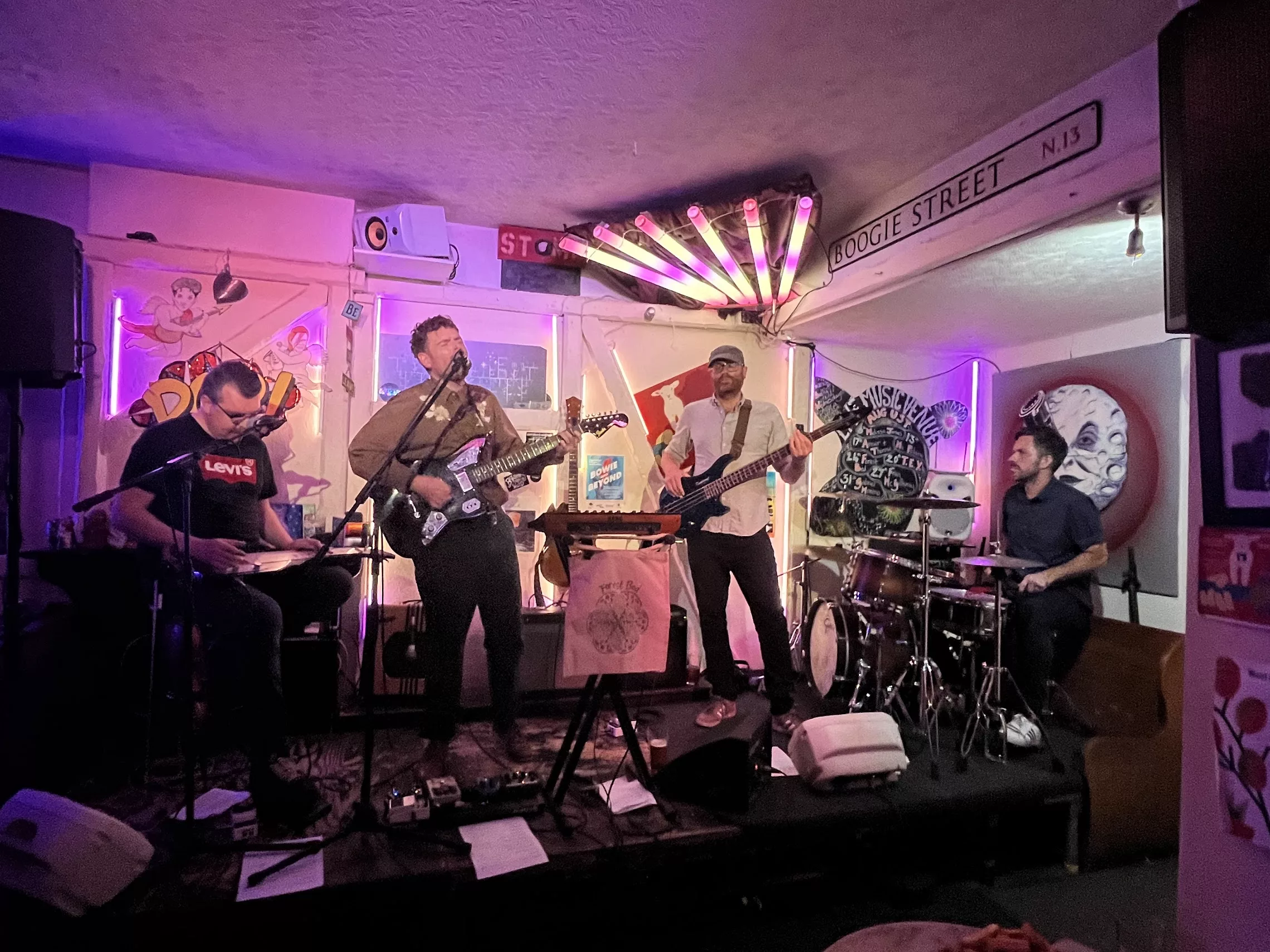 A four-piece band performs on a small, neon-lit stage with a guitarist, bassist, drummer, and pedal steel player under a Boogie Street sign.