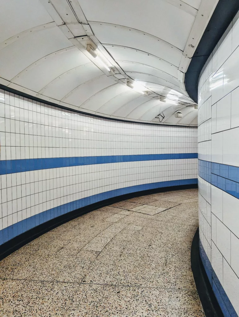 A curved underground station tunnel with white tiles, a horizontal blue stripe, and a vaulted white ceiling with fluorescent lights.