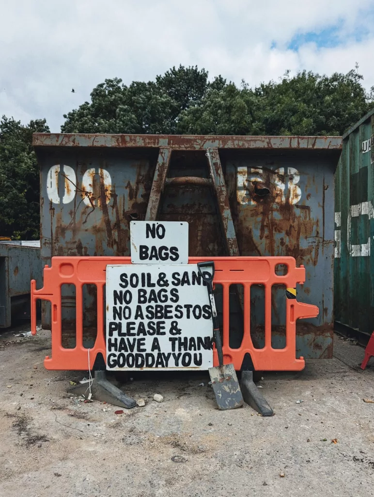A shovel by an orange barrier before a rusty skip. Signs read: NO BAGS and SOIL&SAND NO BAGS NO ASBESTOS PLEASE & HAVE A THANK GOOD DAY YOU.