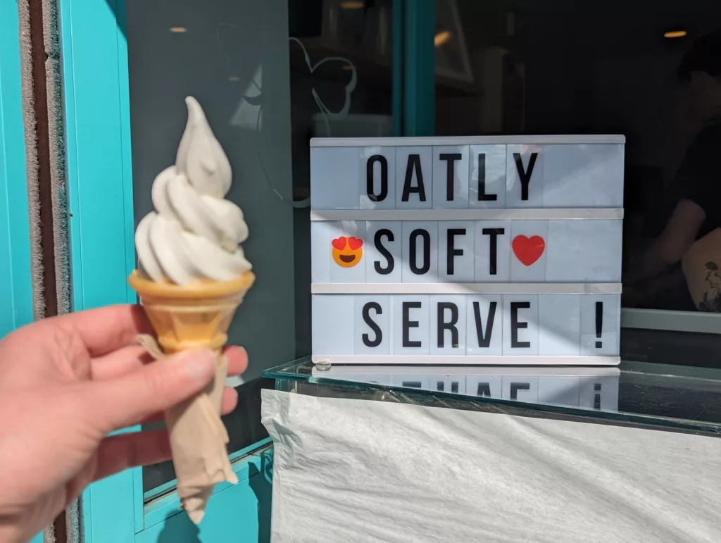 A hand holds a soft serve ice cream cone in front of a light box reading "OATLY SOFT SERVE !" with heart emojis by a bright blue shop frame.