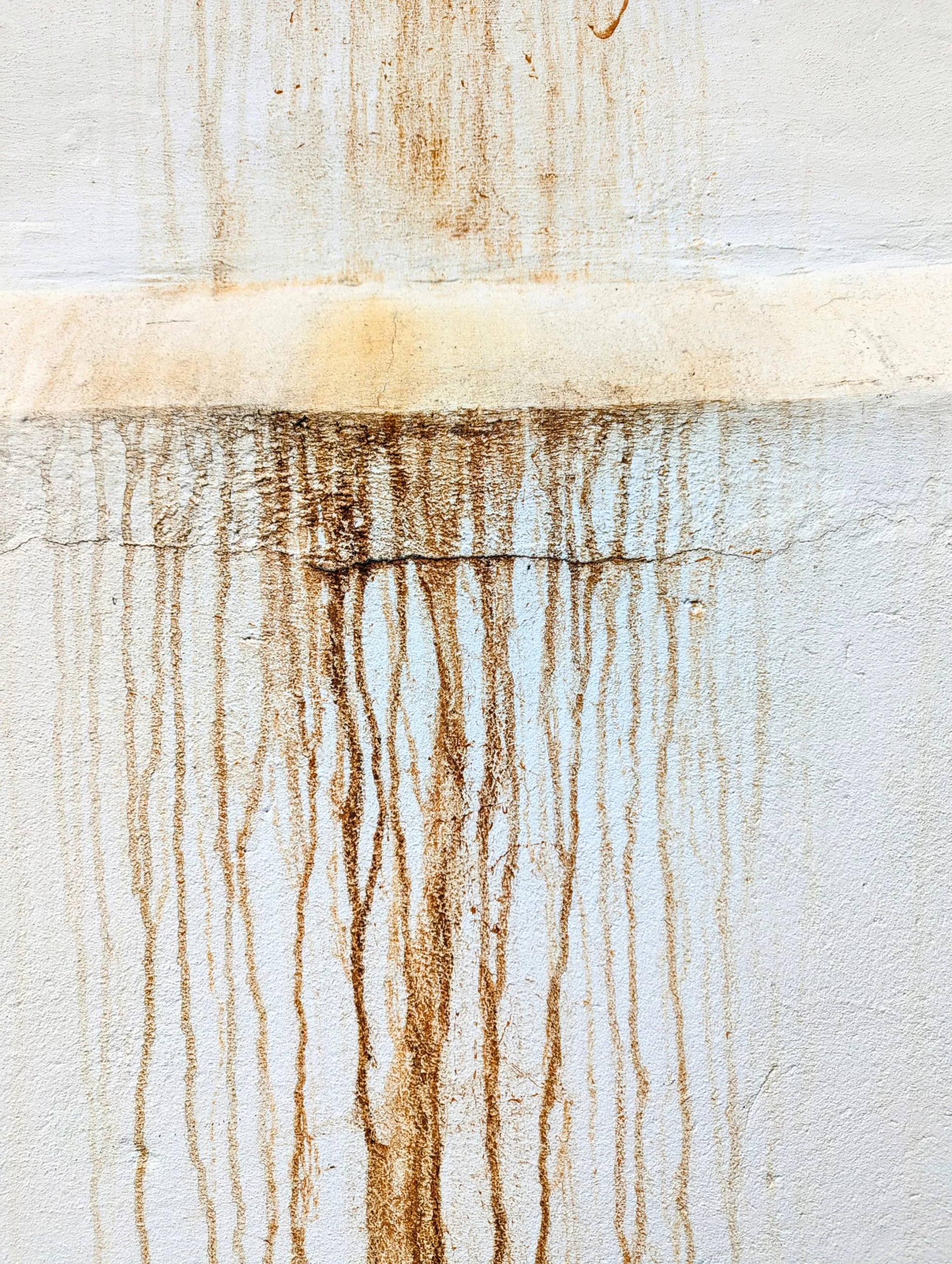 Vertical rust-coloured streaks dripping down a white textured wall from a horizontal ledge and cracks.