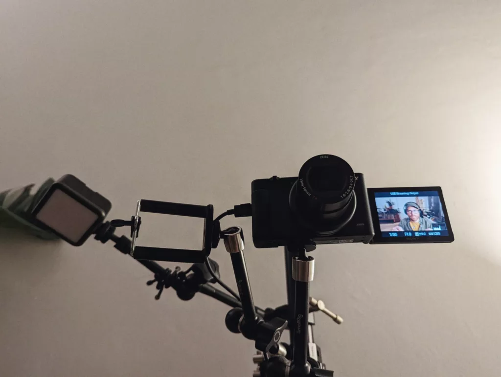 A Sony camera on a tripod rig with a flip-out screen showing a person, a square LED light, and an empty phone mount against a plain white wall.