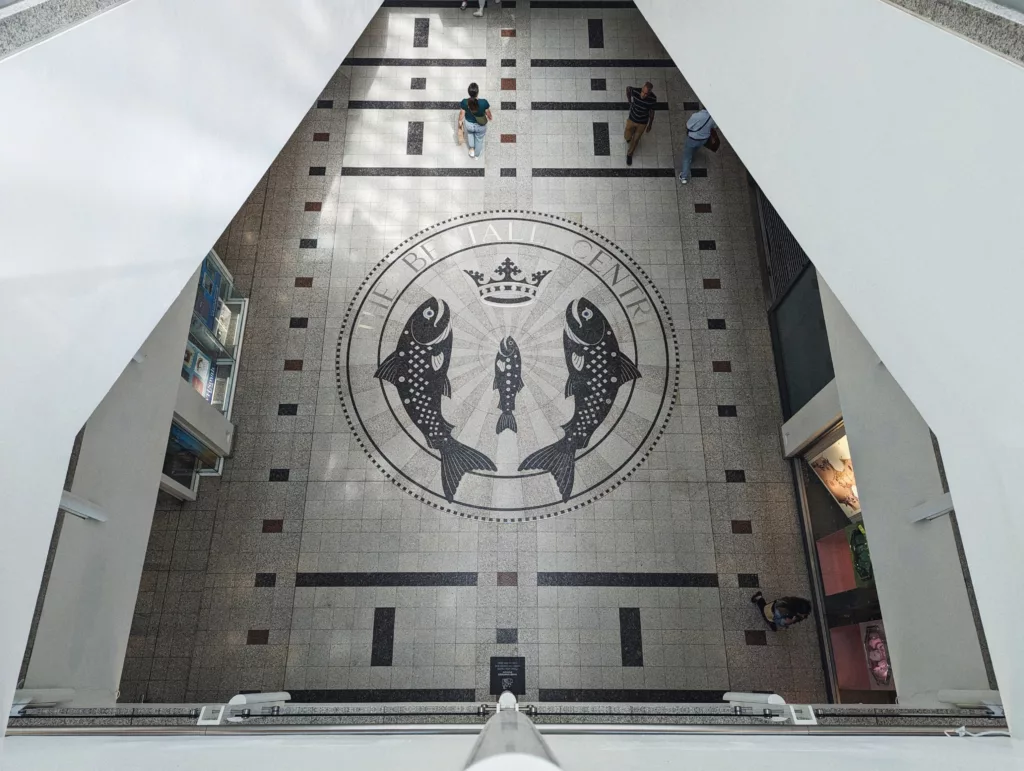 A top-down view of the large fish and crown mosaic on the floor of The Bentall Centre as people walk across the geometric patterned tiles.