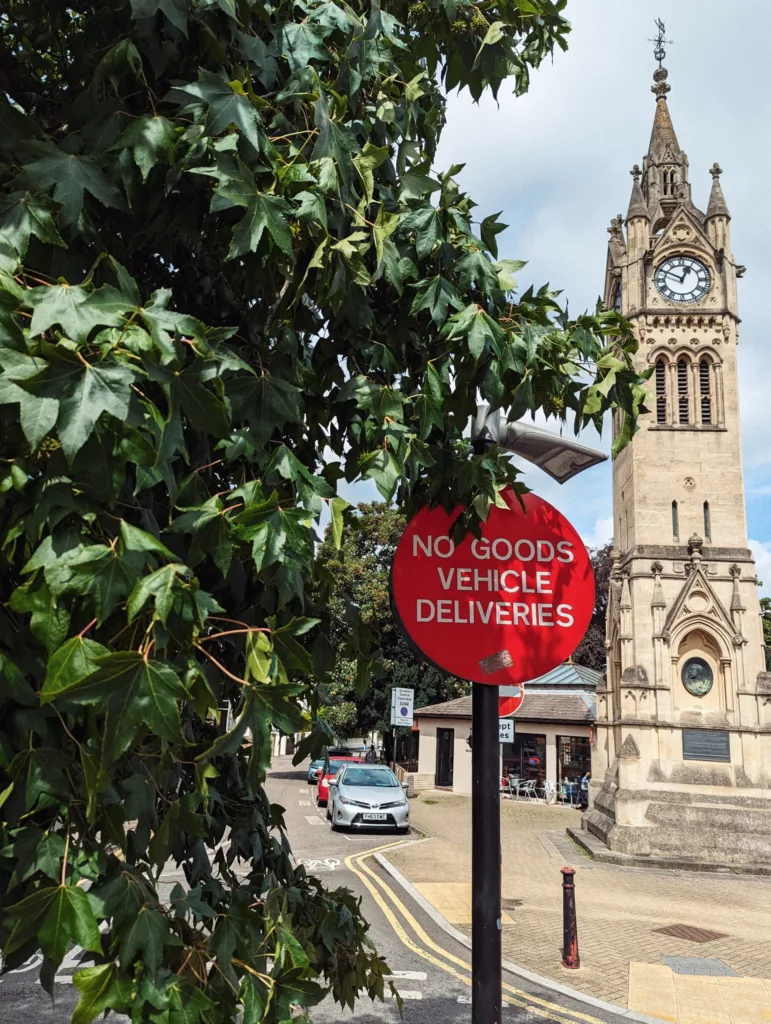 A red sign reading NO GOODS VEHICLE DELIVERIES stands before an ornate stone clock tower, framed by green leaves.