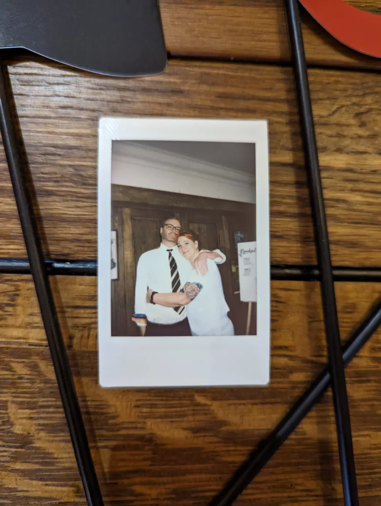 A smiling couple embracing in an instant film photo. The man wears a white shirt and tie; the woman has a rose tattoo on her forearm.