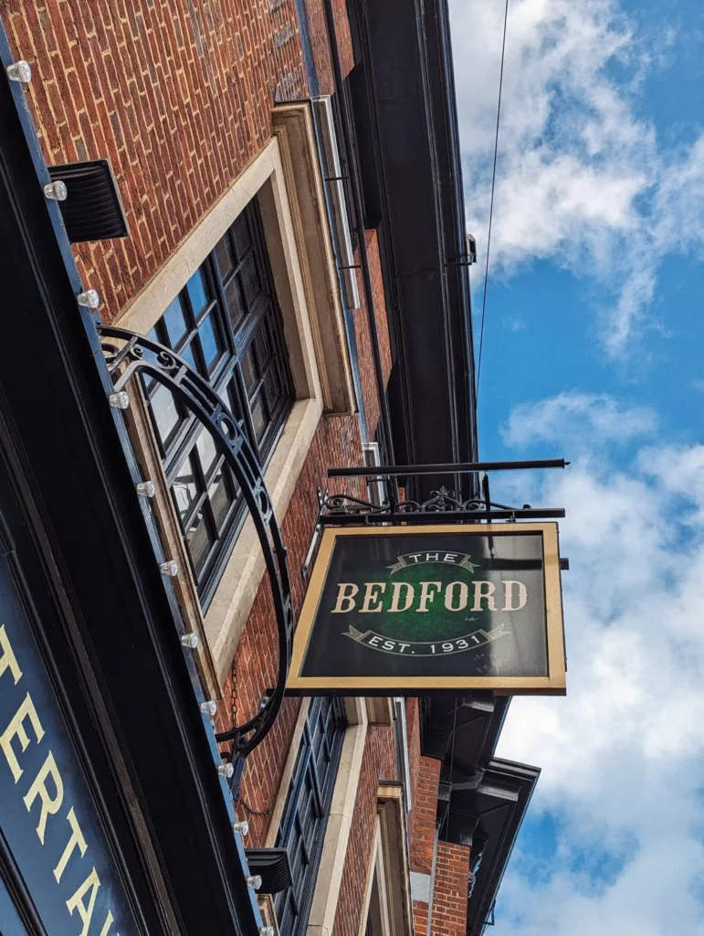 A pub sign for The Bedford Est. 1931 hanging from a brick building facade against a blue sky with white clouds.