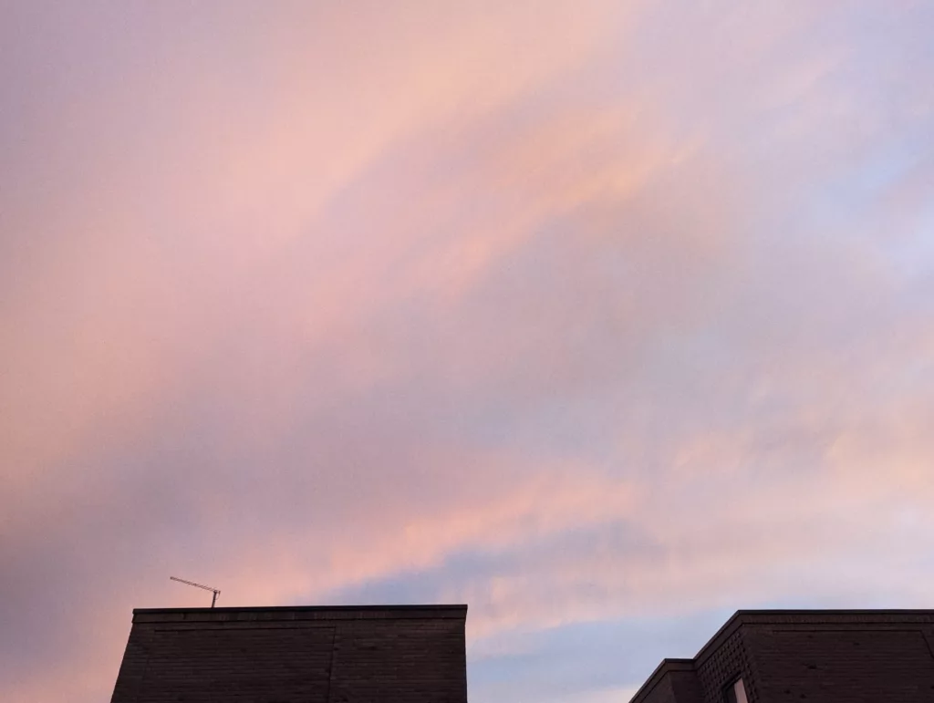 Wispy pink and orange clouds at dusk over the dark silhouettes of two flat-roofed brick buildings.
