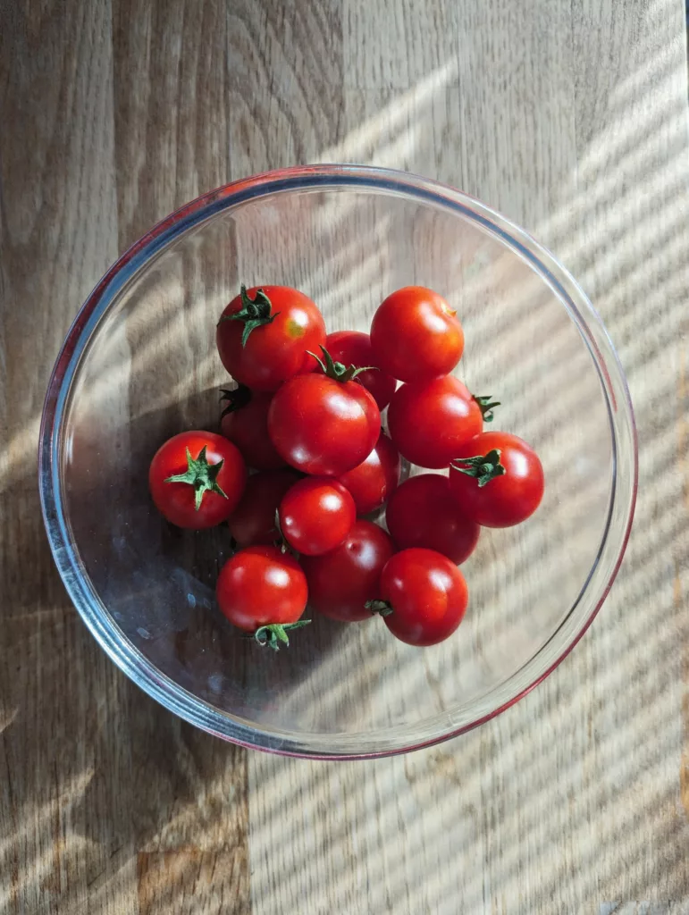 Red cherry tomatoes in a glass bowl on a wooden surface, with sunlight through blinds casting diagonal shadow stripes.