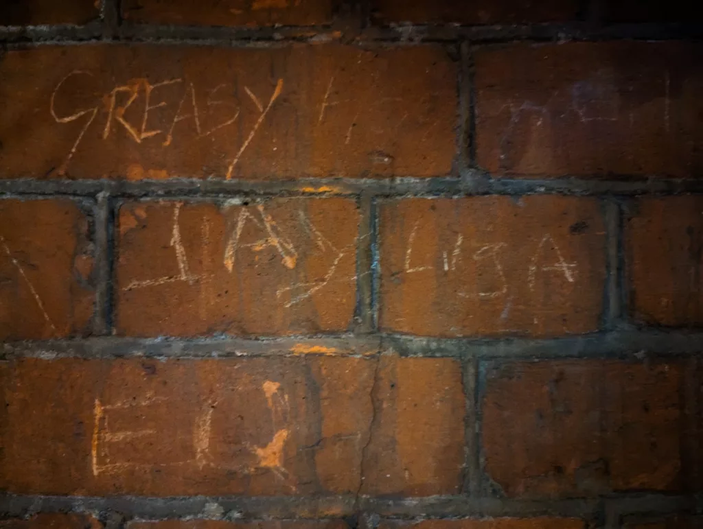 The word GREASY and other graffiti roughly scratched into the surface of dark, weathered orange bricks in dim lighting.