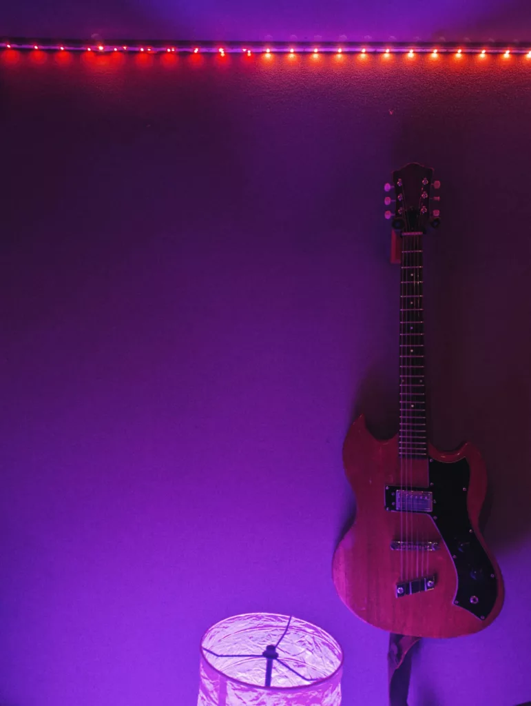 An electric guitar hanging on a wall bathed in deep purple light, with a red LED strip at the ceiling and a glowing lamp shade in the foreground.