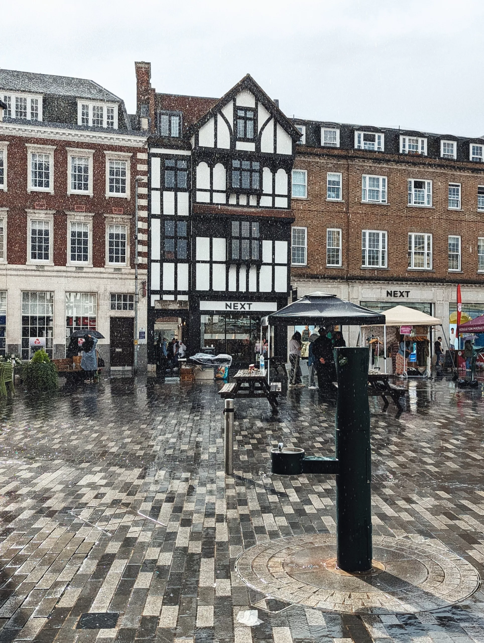 Rain falling on a wet paved market square in Kingston, featuring a black water fountain and a distinctive Tudor-style Next storefront.