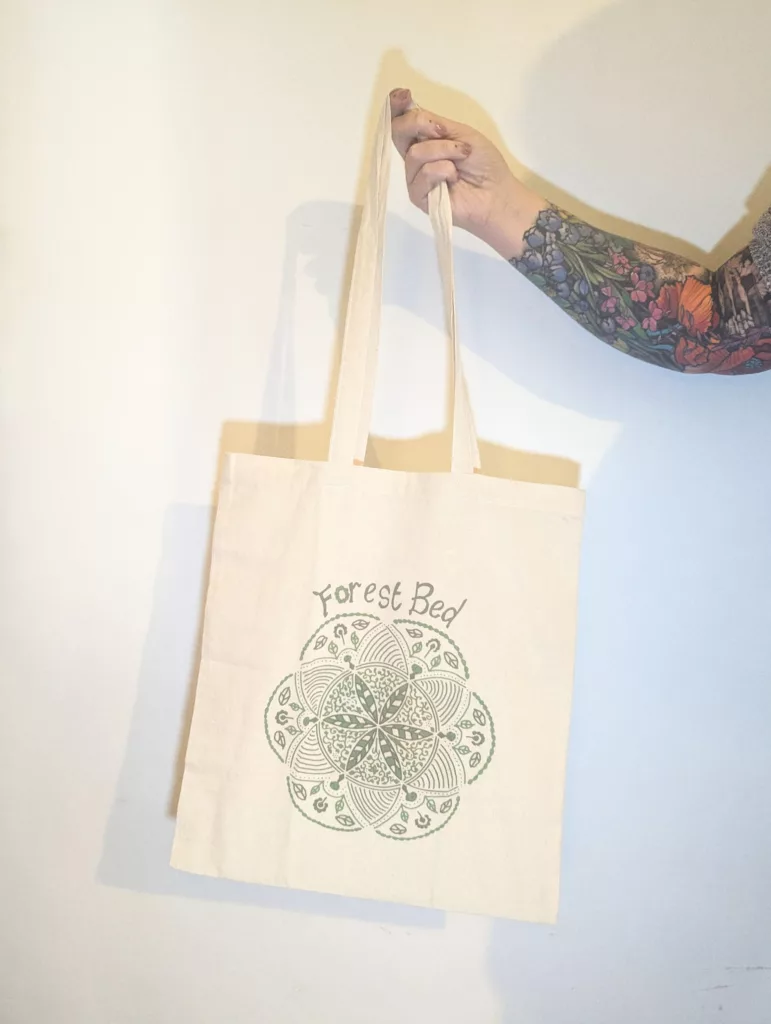 A hand with a floral tattoo holding a cream tote bag printed with Forest Bed and a green mandala design against a plain wall.