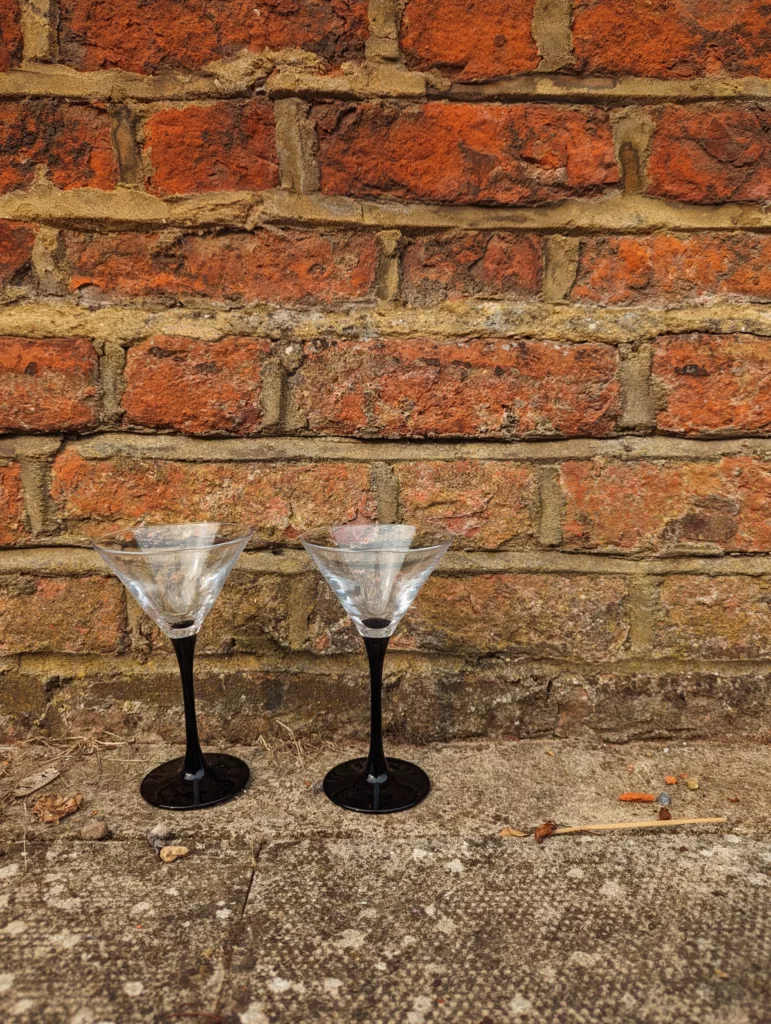 Two martini glasses with black stems and clear bowls sit on a concrete surface against a weathered red brick wall.