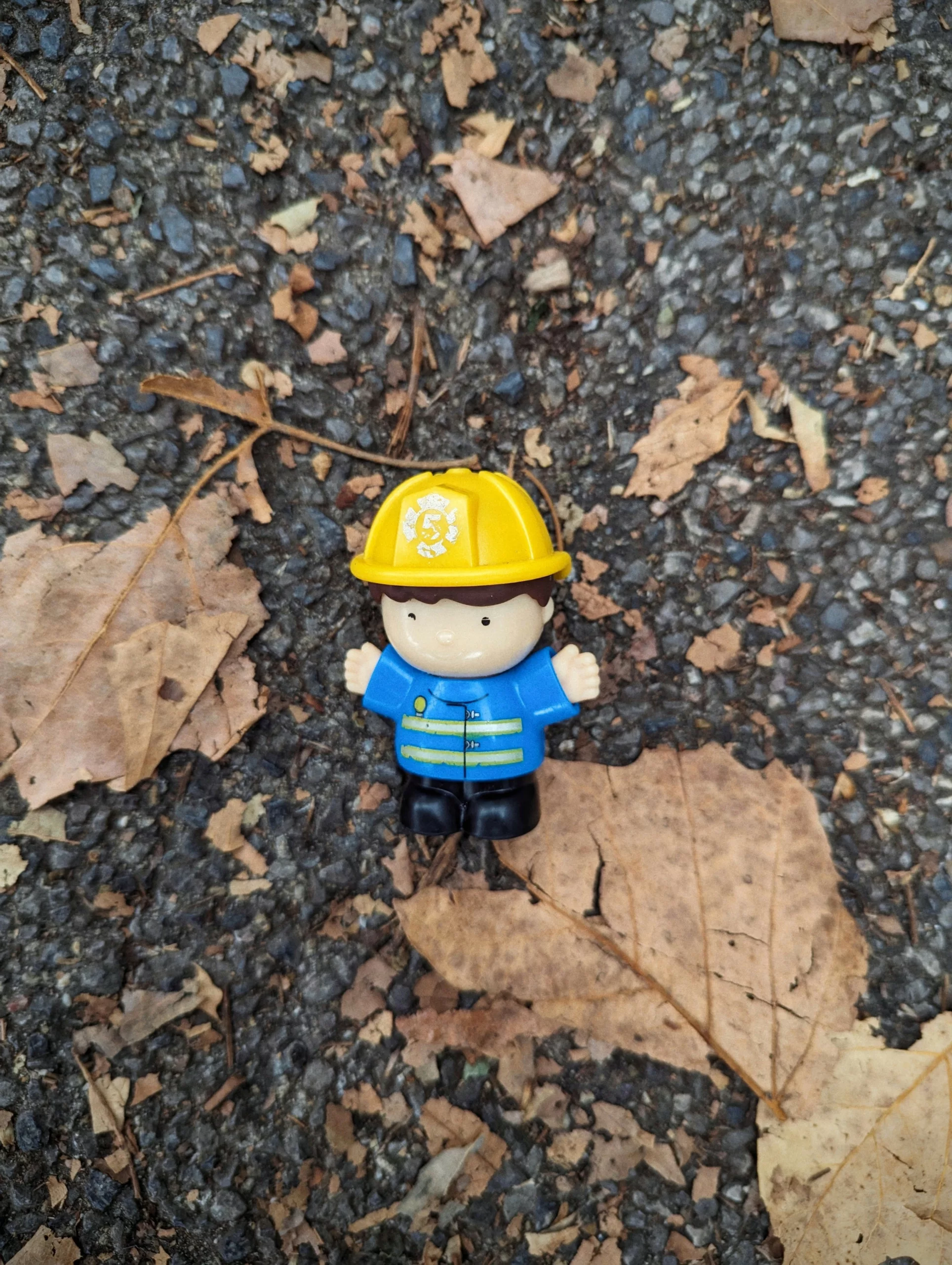 A small toy firefighter with a yellow helmet and blue jacket on an asphalt ground covered in dry autumn leaves.