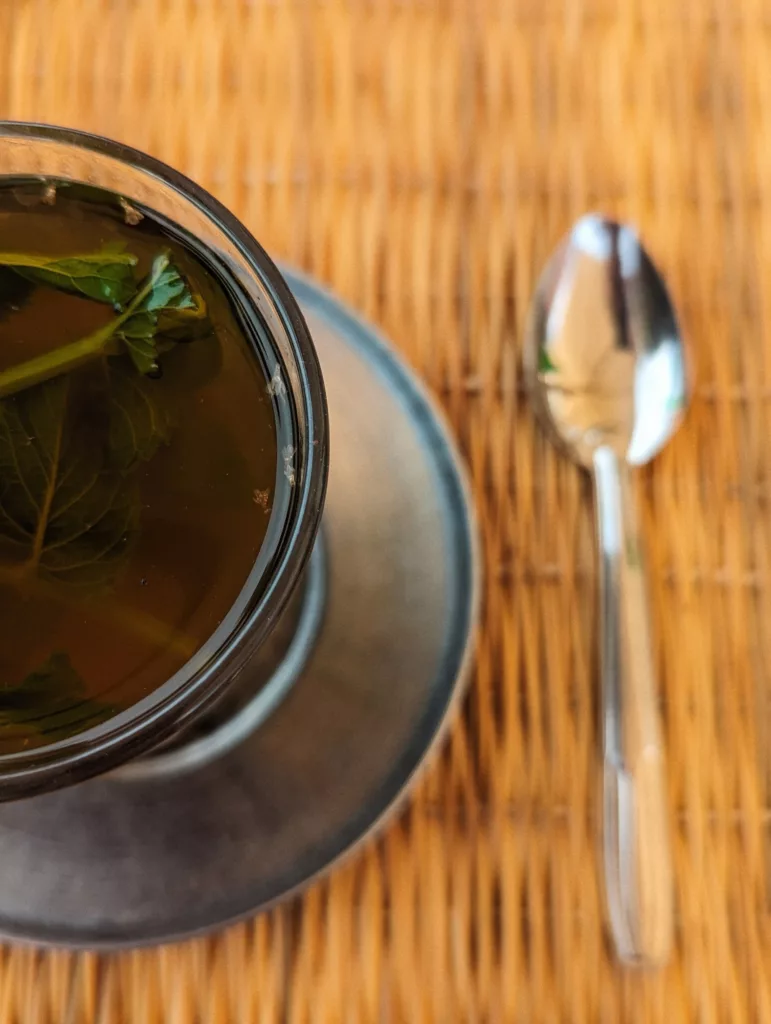 Fresh mint tea in a clear glass on a silver saucer next to a teaspoon, resting on a brown woven mat.