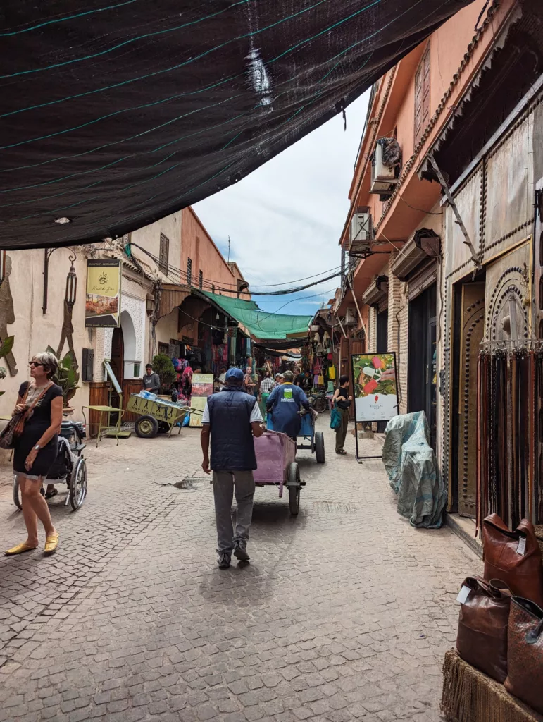 A man pushes a purple cart through a narrow street in a Moroccan medina, with terracotta buildings and shop displays under overhead shade cloths.