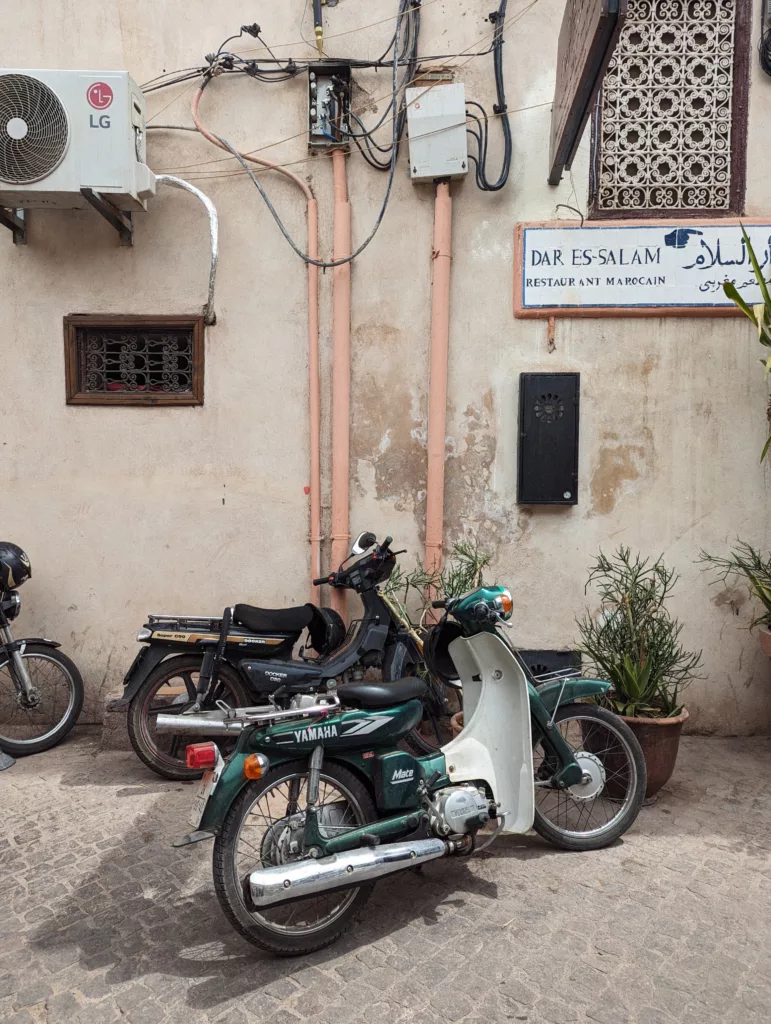 A green Yamaha Mate moped parked on a cobblestone street by a weathered wall with exposed wiring and a sign for a Moroccan restaurant.