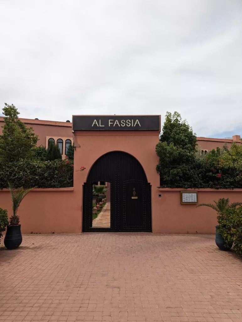 A pink terracotta entrance with a black arched gate and a sign for AL FASSIA, surrounded by green trees and bushes.