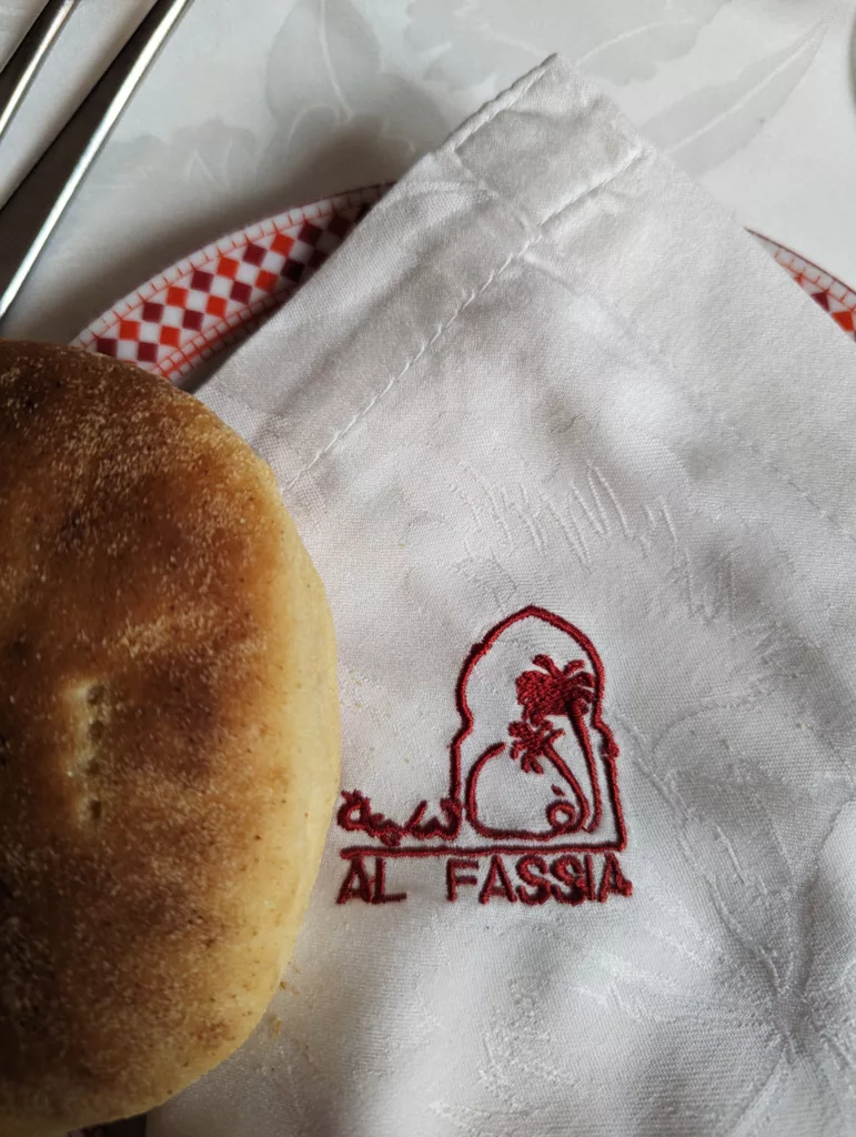 Close-up of khobz bread beside a white linen napkin embroidered with the red Al Fassia restaurant logo.