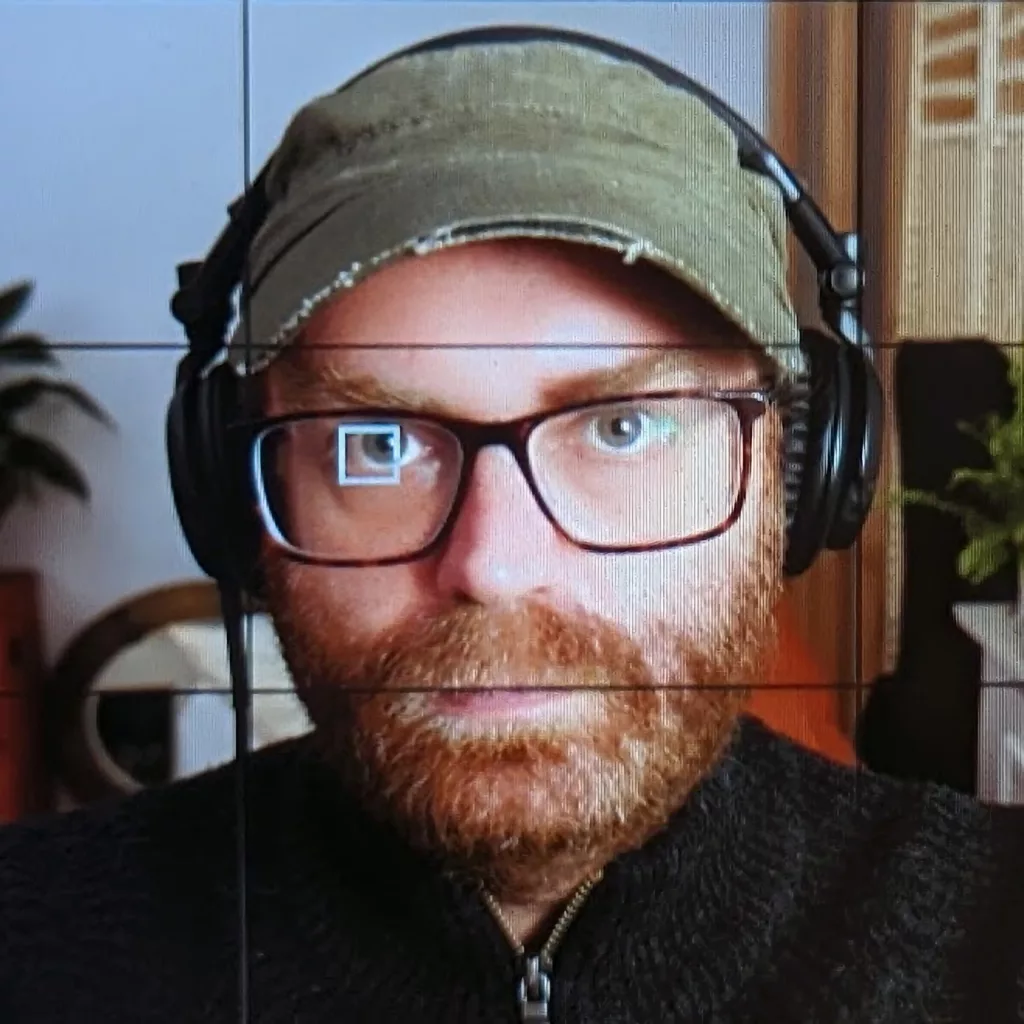 A man with a ginger beard, green distressed cap, and glasses wearing black headphones, shown with a camera focus square and grid overlay.